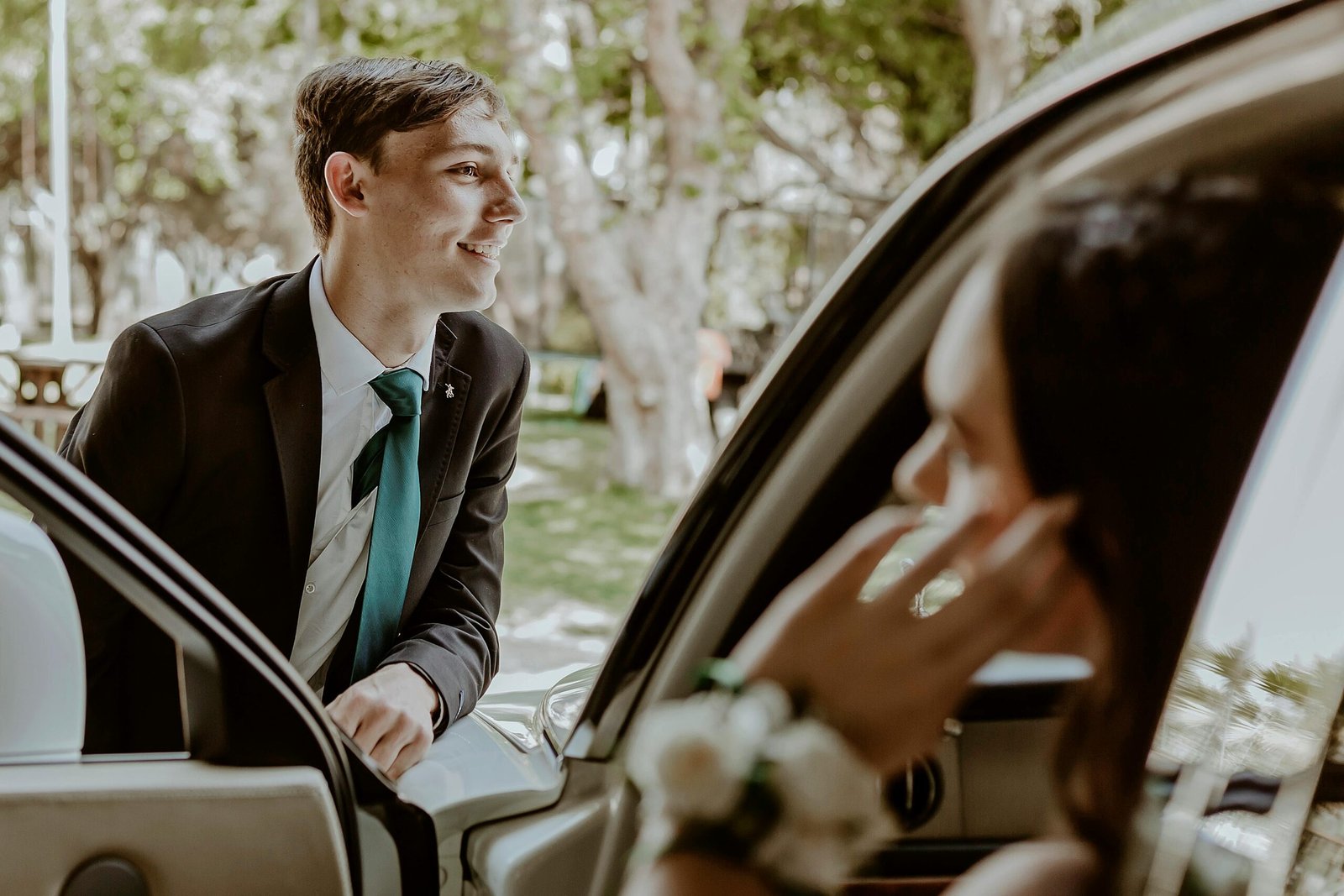 A young couple enjoying a sunny day in formal attire, smiling by a car outdoors.
